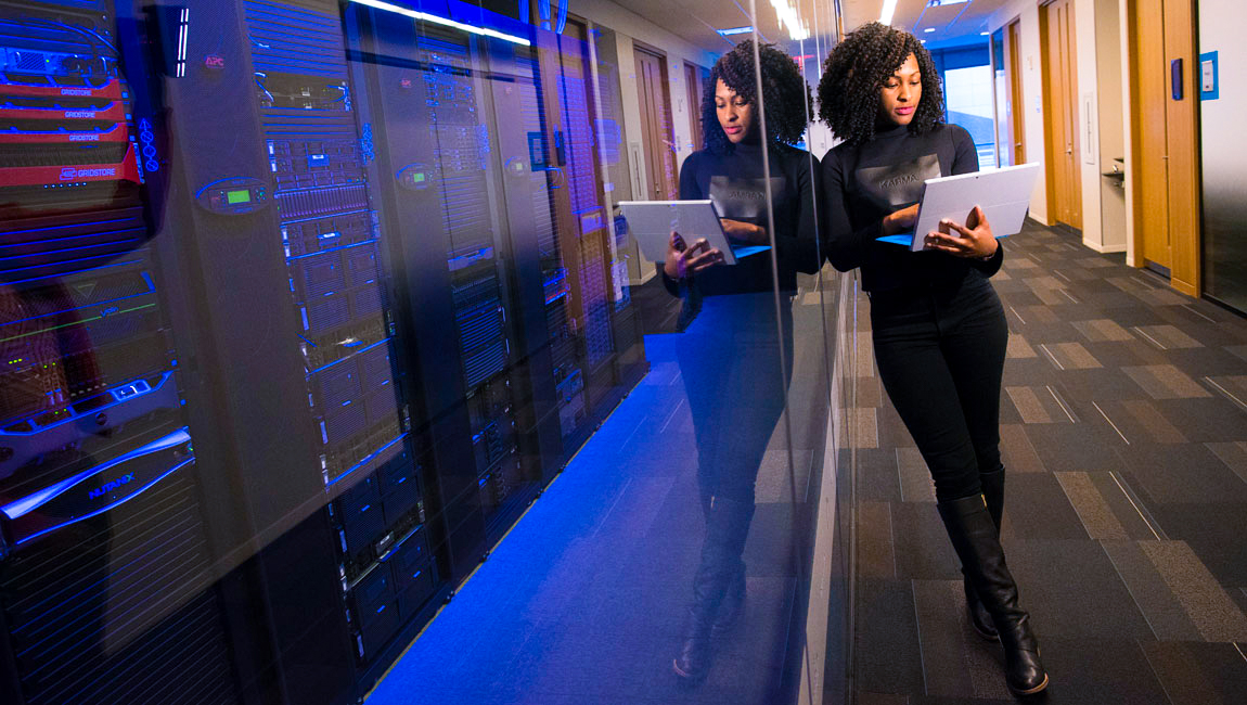 Business woman leaning against wall with computer servers looking at her laptop